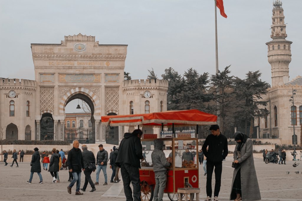People walk near İstanbul University's grand entrance, with a street vendor in the foreground.