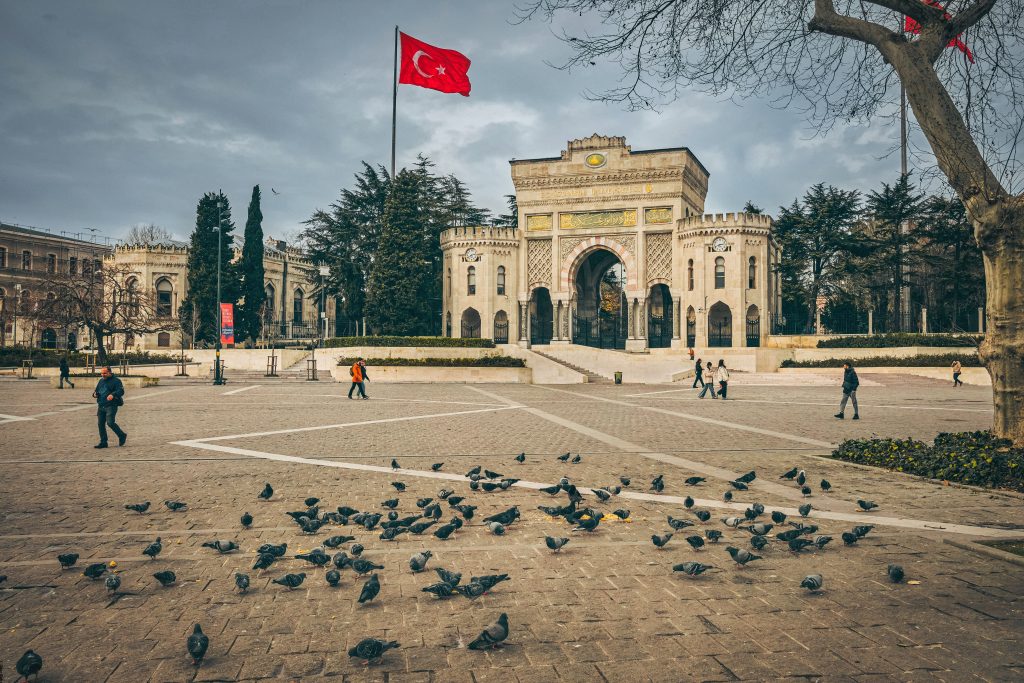 Istanbul University gate with pigeons and Turkish flag in a bustling city square.